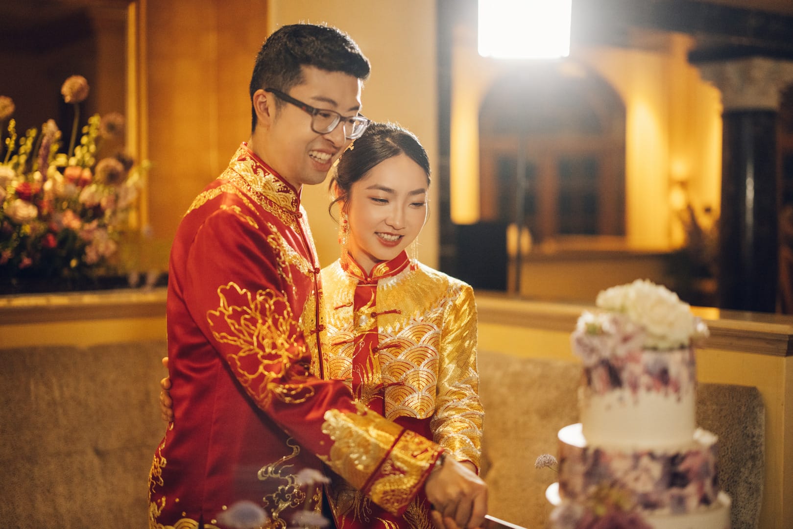 A front image of a bride wearing a traditional Chinese Wedding Dress with a gold embroidered lucky seawaves jacket with her groom in matching red tang suit at their Chinese tea ceremony.