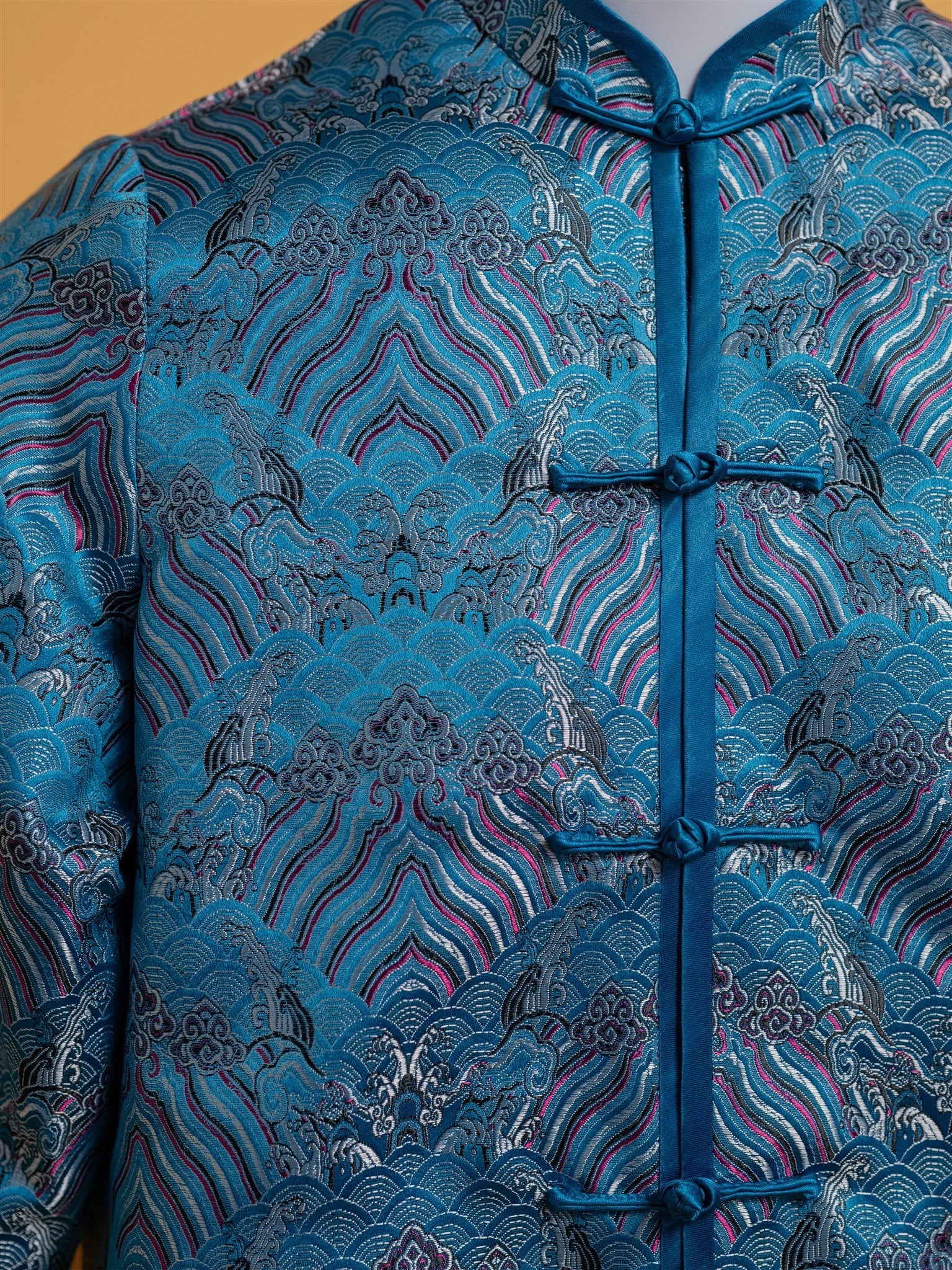 Close-up detail of the frog knot button and intricate brocade pattern on a blue silk Tang suit with a mandarin collar.