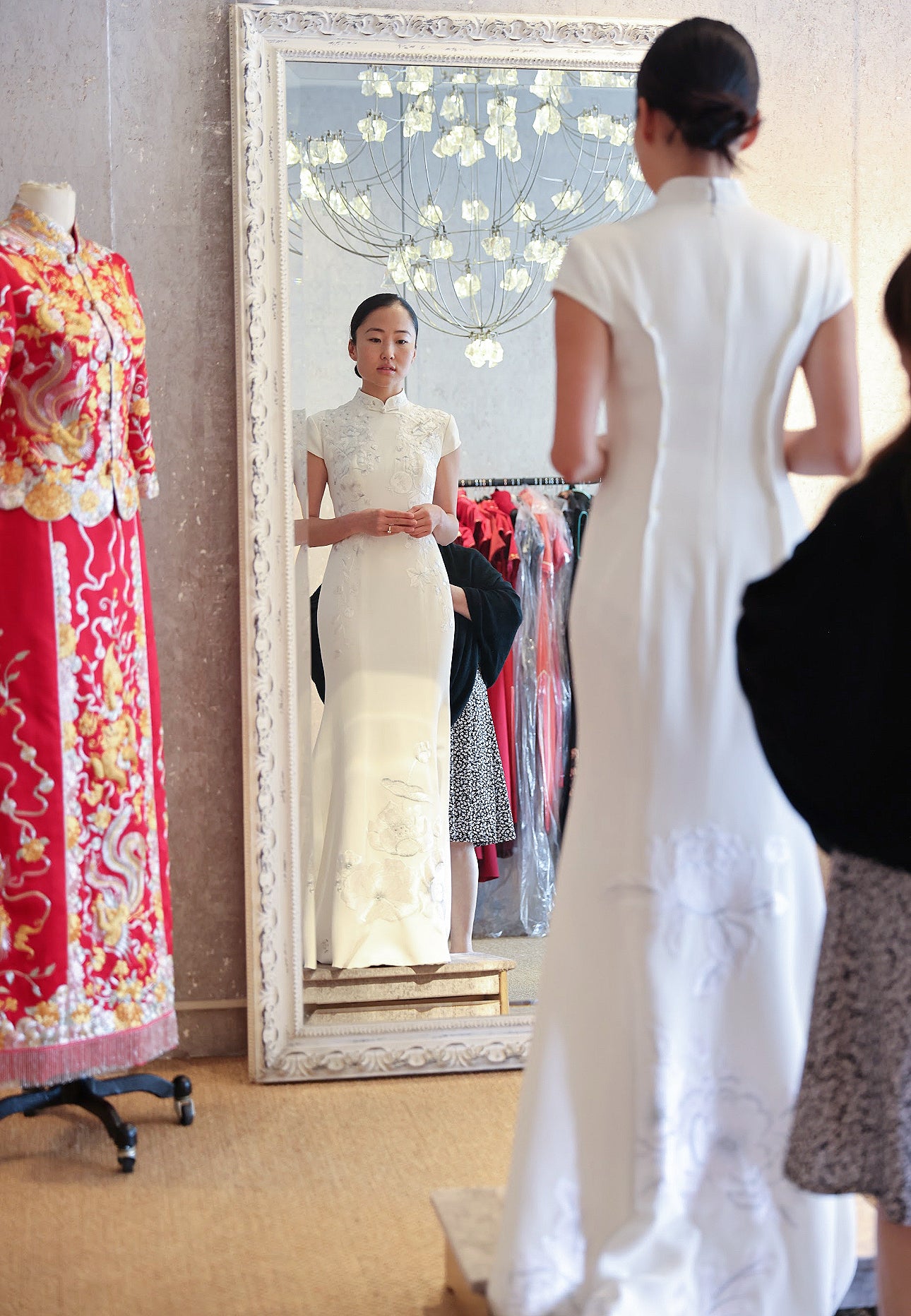 Bride Connie in a white silk crepe modern Qipao with sleeves and a fitted-flare silhouette, featuring white and silver embroidery of peonies, pomegranates, and lotus flowers, celebrating her Jewish and Chinese heritage.